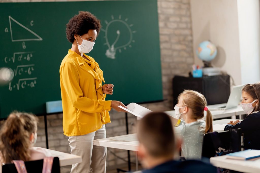 Happy black teacher giving exam paper to her student while wearing protective face mask in the classroom